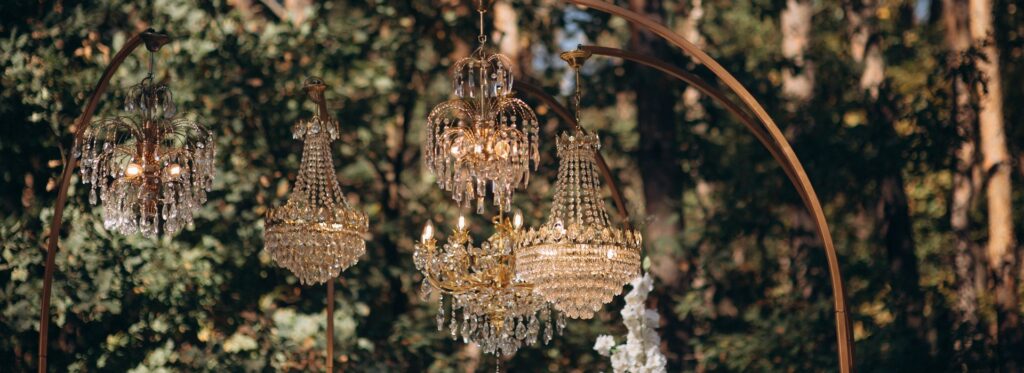 Crystal chandeliers hanging from a gold arch at an outdoor woodland wedding